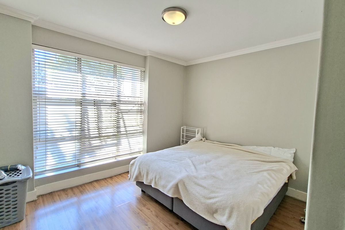 A bright bedroom featuring laminate flooring, crown molding, and large windows fitted with venetian blinds, providing a neutral and clean living space.