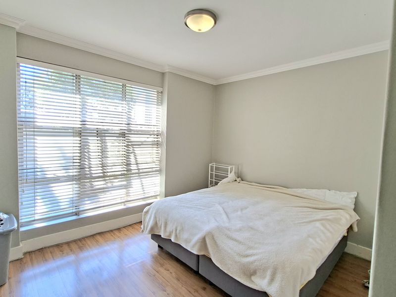 A bright bedroom featuring laminate flooring, crown molding, and large windows fitted with venetian blinds, providing a neutral and clean living space.