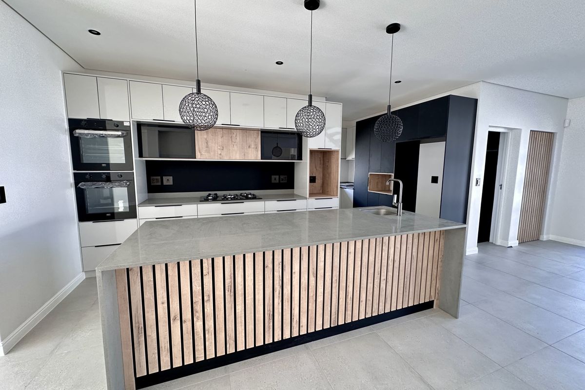 Modern kitchen featuring a large central island with stone countertop, integrated double ovens, gas hob, and contemporary cabinetry with wood-slat detailing.