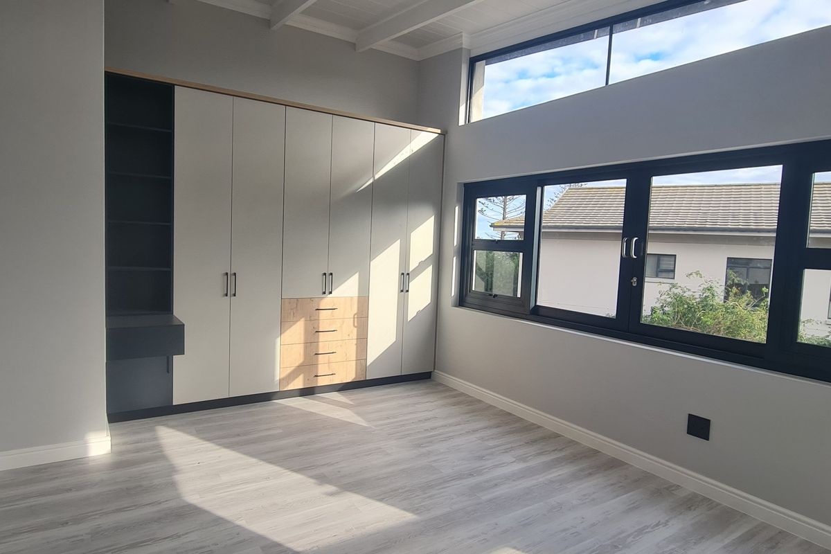 A bright, modern bedroom featuring built-in cupboards, light laminate flooring, and large aluminium windows with clerestory panels for natural light.