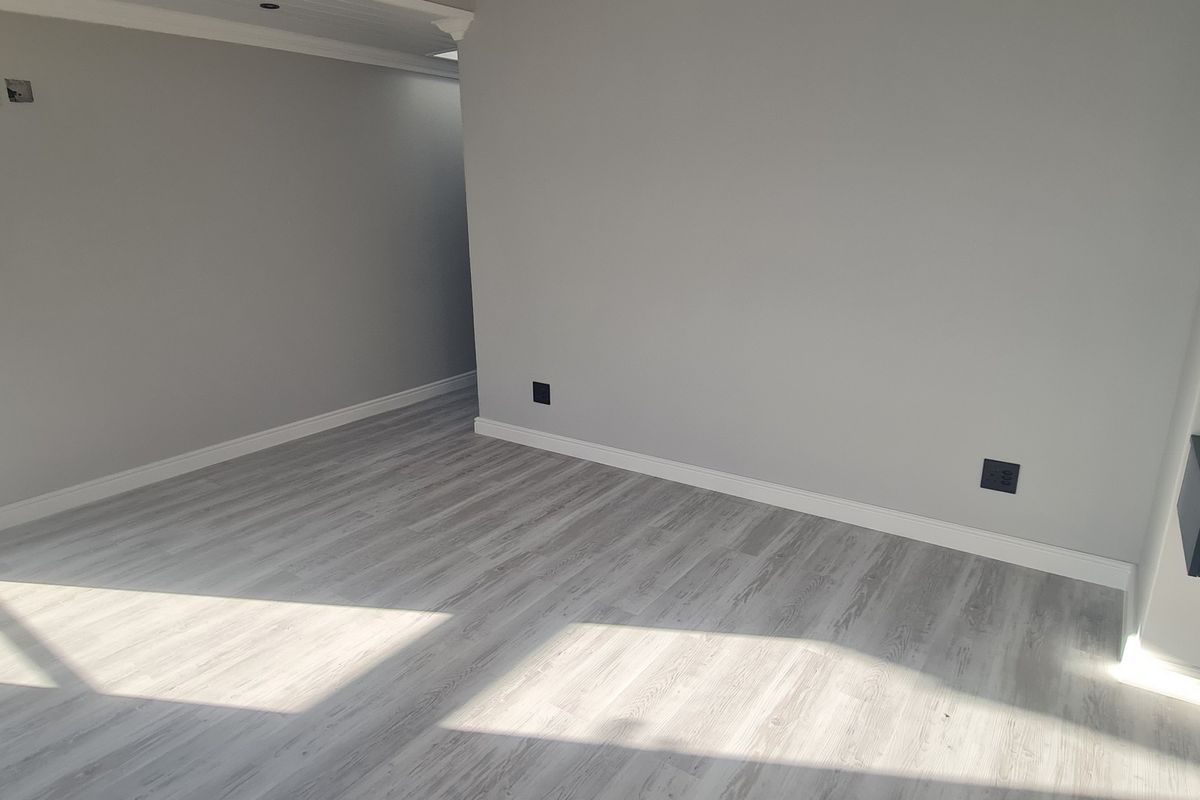 A bright, empty living area featuring modern light-toned laminate flooring, neutral grey walls, and a sleek floating shelf unit.