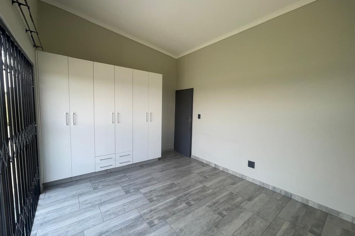 A clean, empty bedroom featuring modern grey wood-look floor tiles, neutral walls, and ample built-in cupboard storage with a secure entrance gate.
