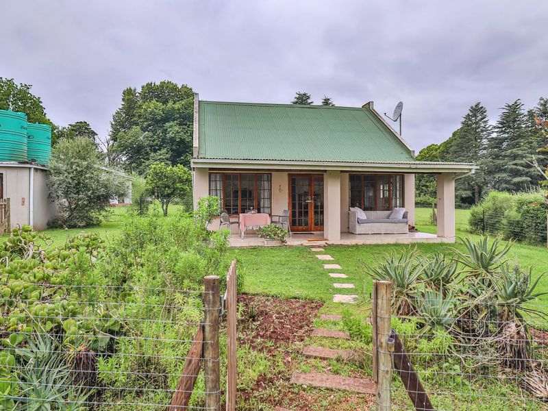 A charming cottage featuring a green corrugated metal roof, a covered patio, and external water storage tanks set within a spacious garden.