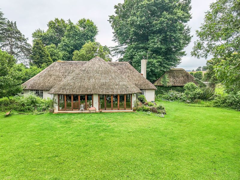 A rear view of a charming thatched-roof house featuring expansive timber-framed folding doors opening onto a large, well-maintained green lawn and mature garden.