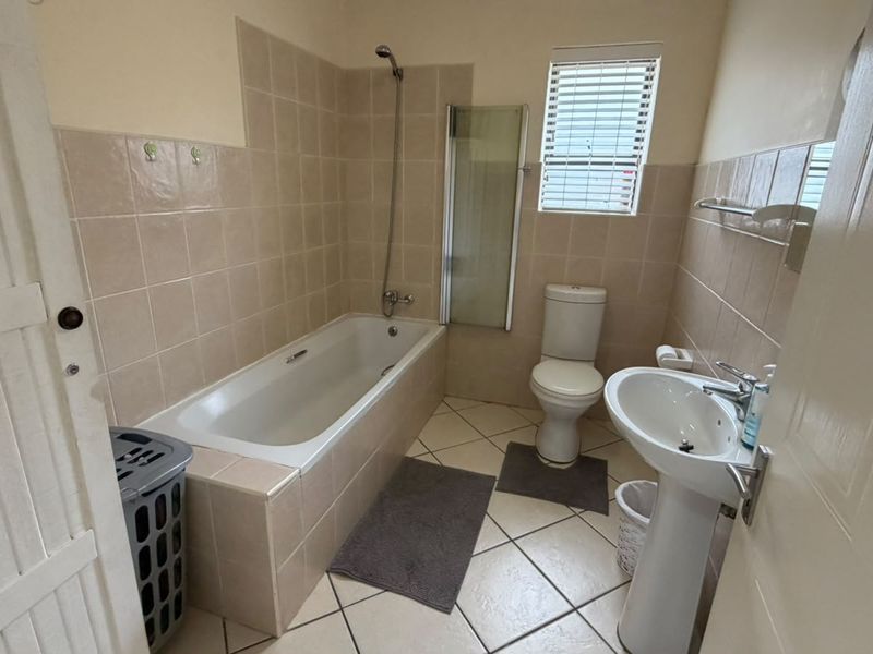 A functional bathroom featuring a bathtub with a shower head, glass screen, pedestal sink, and neutral wall and floor tiling.