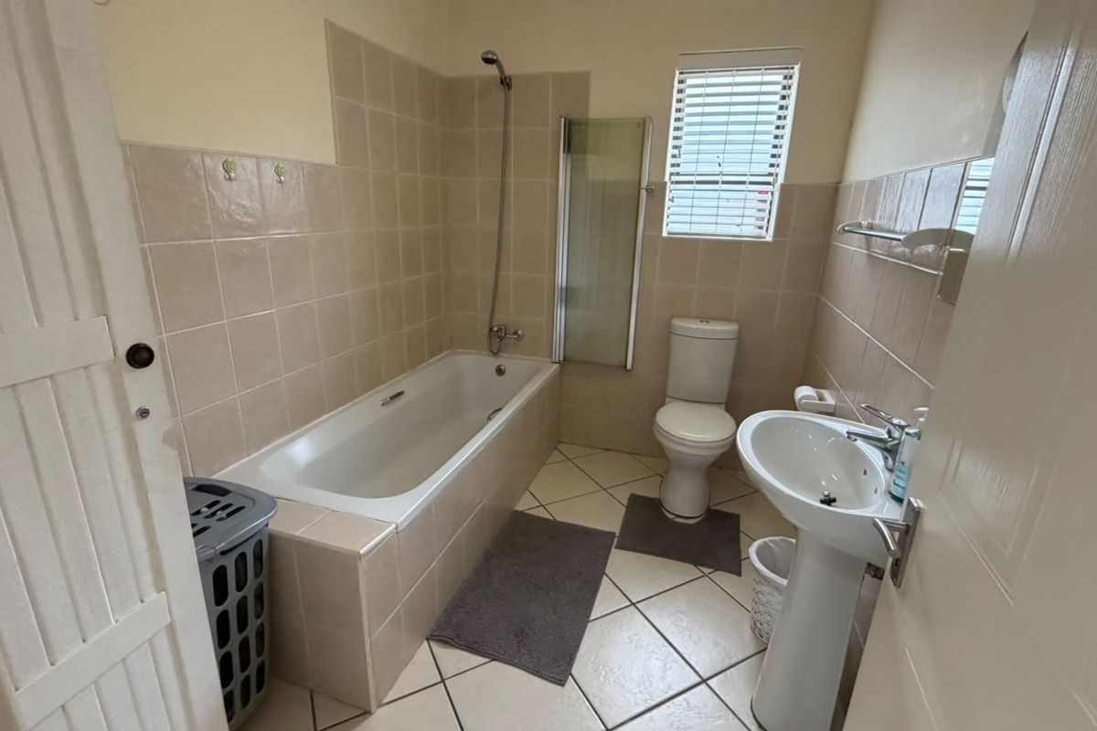 A functional bathroom featuring a bathtub with a shower head, glass screen, pedestal sink, and neutral wall and floor tiling.