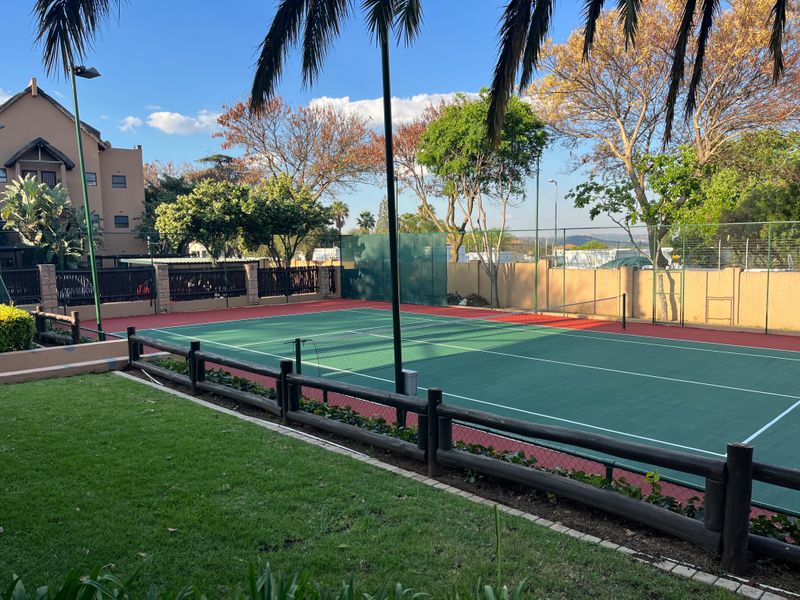 A well-maintained outdoor tennis court featuring a green surface, perimeter fencing, and floodlights, set within a landscaped residential complex.