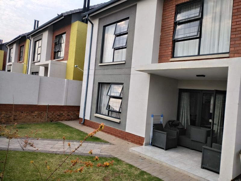 Rear view of a modern townhouse featuring a covered tiled patio, paved garden walkway, and well-maintained brick and plaster facade with aluminium windows.