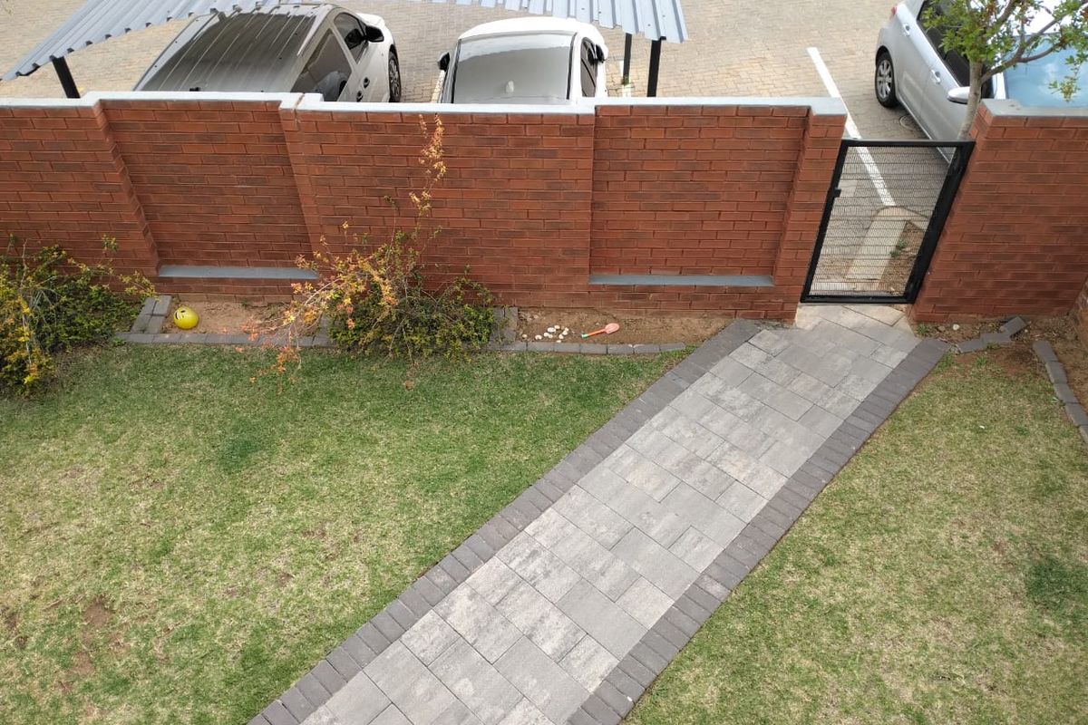 An elevated view of a private rear garden featuring a paved walkway, manicured lawn, and a brick boundary wall with a pedestrian gate.
