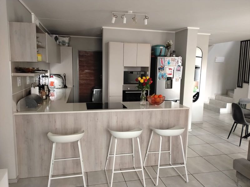 Modern open-plan kitchen featuring light wood-grain cabinetry, stone countertops, integrated appliances, and a breakfast bar, seamlessly flowing into the living and staircase area.