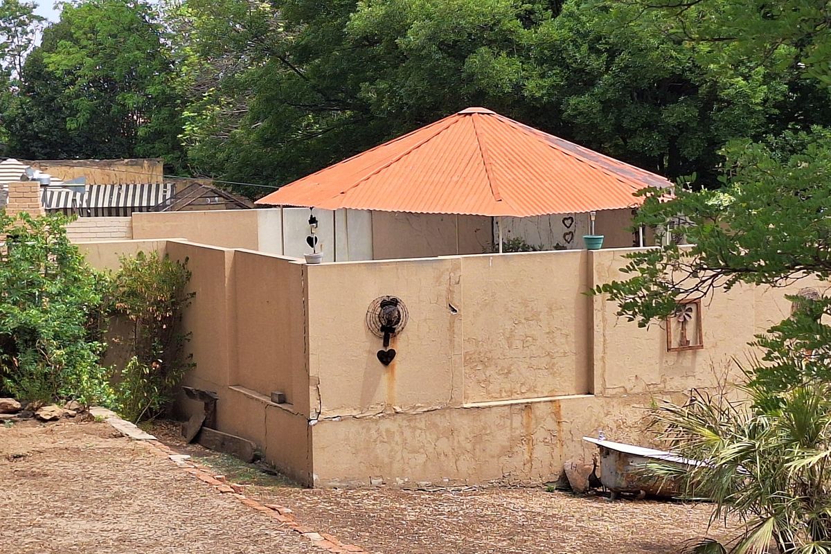 An outdoor structure with a corrugated metal roof and plastered walls, showing signs of significant weathering and maintenance requirements in a garden setting.