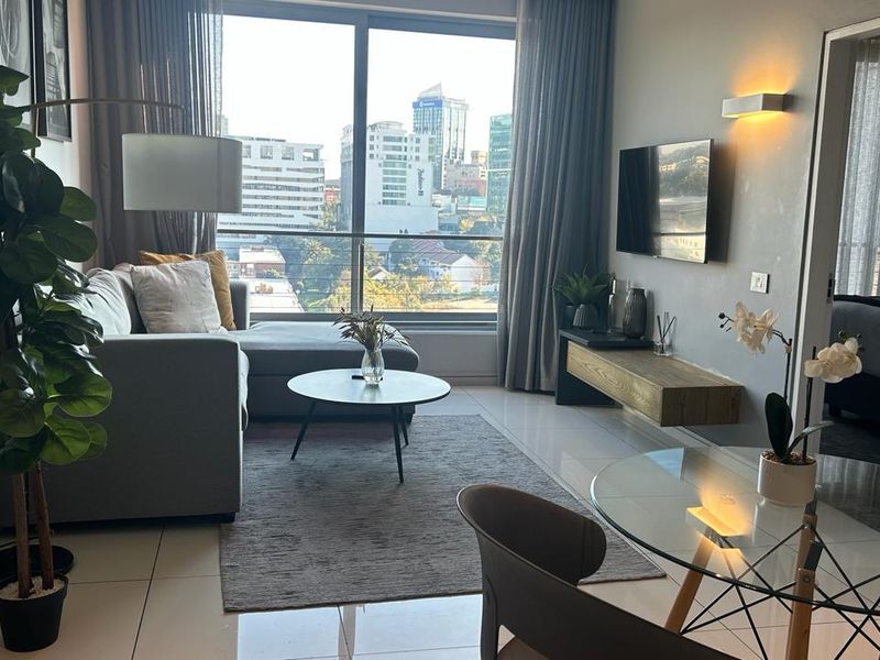 Modern open-plan living area featuring large floor-to-ceiling windows with city views, tiled flooring, and a sleek wall-mounted media console.