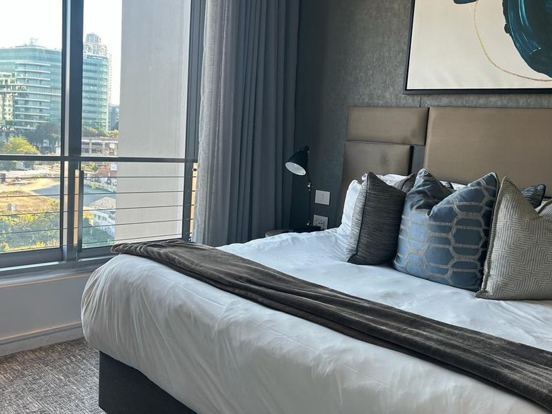 A well-appointed bedroom featuring a large upholstered headboard, carpeted flooring, and floor-to-ceiling windows offering an urban view.