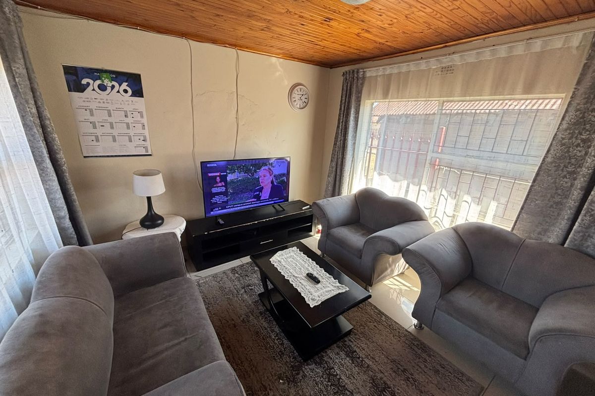 A living room featuring a timber tongue and groove ceiling, tiled flooring, and a large window, showing signs of wear on the walls.