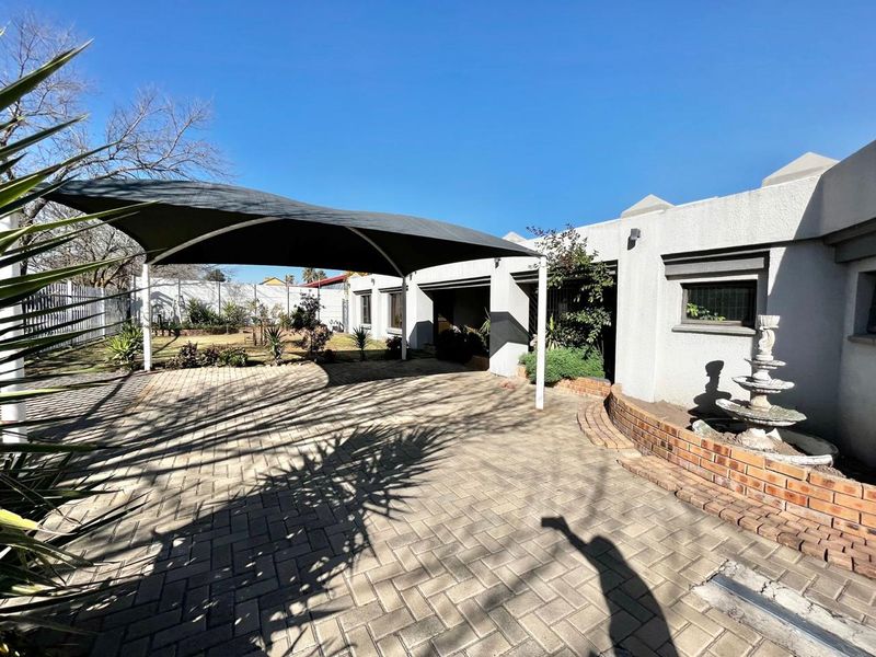 Rear exterior view featuring a paved courtyard, a large shade sail structure, a brick retaining wall, and a decorative stone fountain.