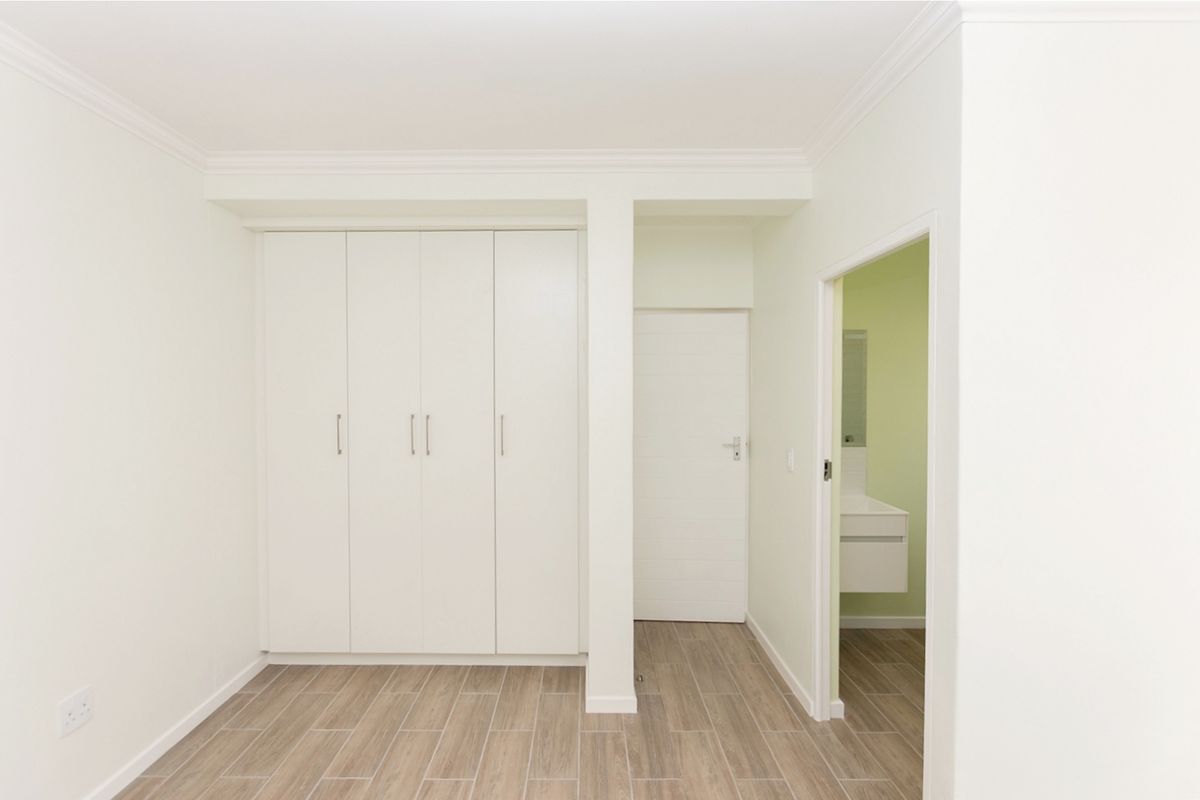 A clean, empty bedroom featuring built-in cupboards, modern wood-look tiled flooring, and neutral wall finishes with access to an adjoining bathroom.