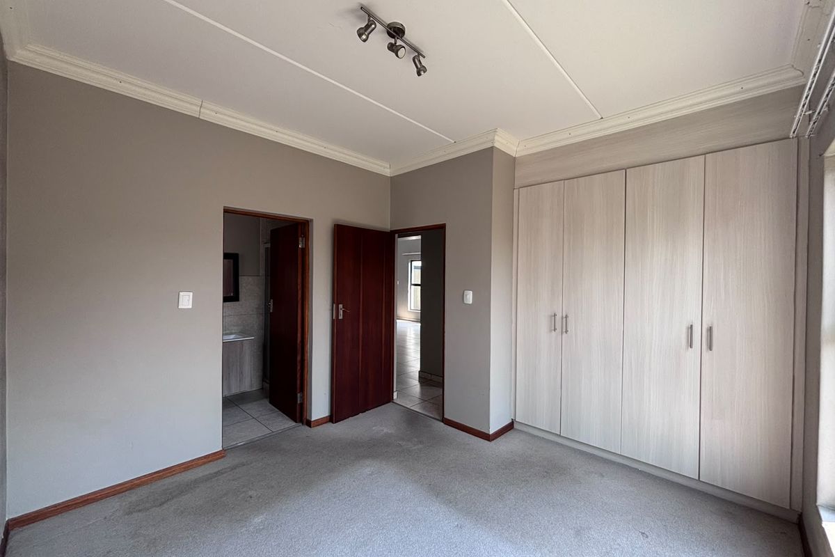 A spacious, empty bedroom featuring neutral grey walls, built-in cupboards with light wood-grain finish, and carpeted flooring, with direct access to an en-suite bathroom.