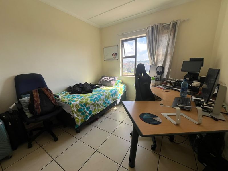 A functional bedroom featuring tiled flooring and a window with natural light, currently utilized as a combined sleeping and workspace area.