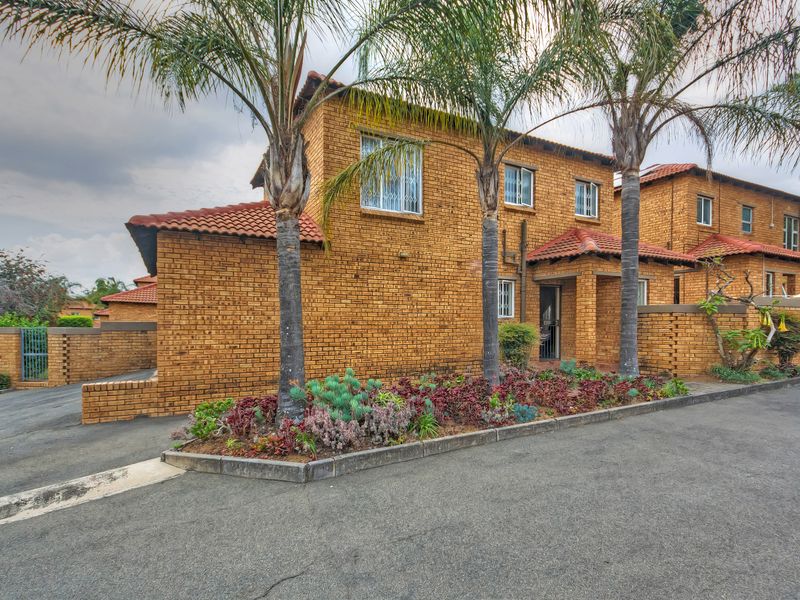 A well-maintained double-story townhouse featuring a classic face brick exterior, terracotta roof tiles, and a neat front garden with mature palm trees.