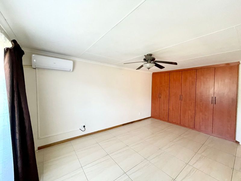 A spacious bedroom featuring neutral tiled flooring, built-in wooden cupboards, a ceiling fan, and a wall-mounted air conditioning unit.