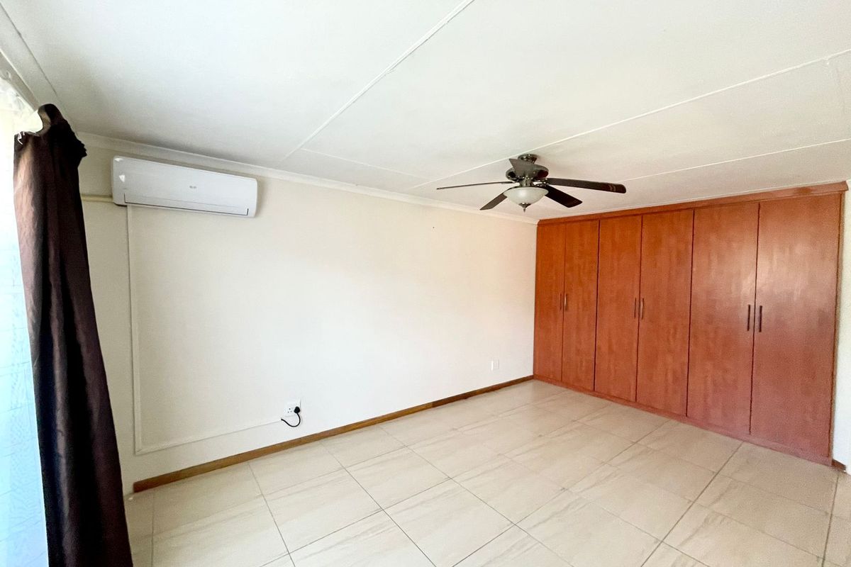 A spacious bedroom featuring neutral tiled flooring, built-in wooden cupboards, a ceiling fan, and a wall-mounted air conditioning unit.