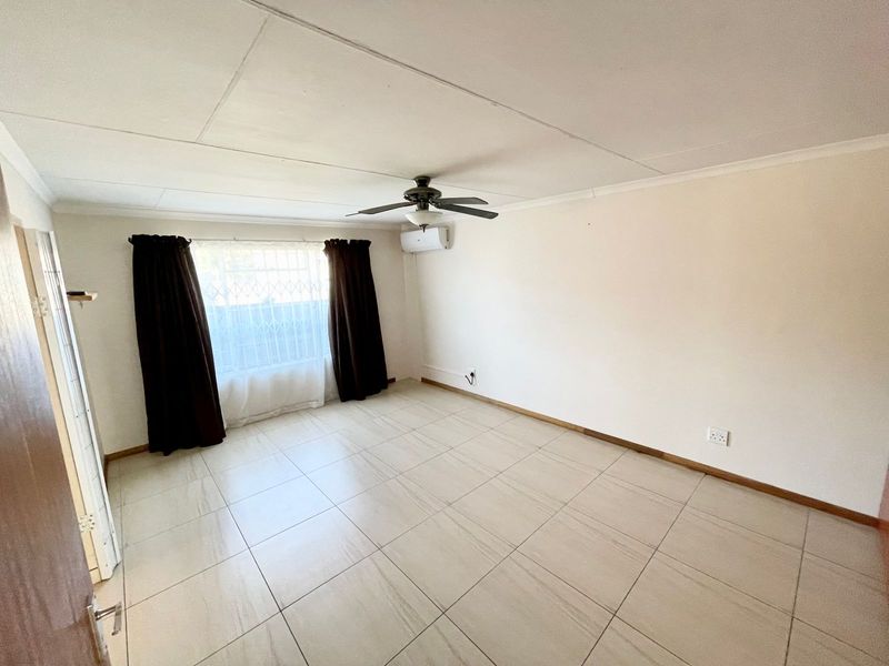A bright, empty living room featuring neutral tiled flooring, a ceiling fan, and a wall-mounted air conditioning unit.