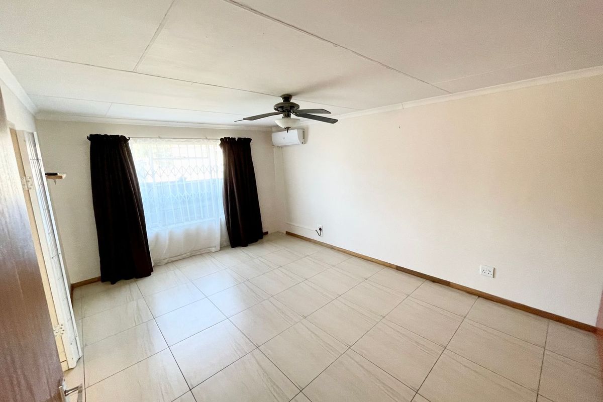 A bright, empty living room featuring neutral tiled flooring, a ceiling fan, and a wall-mounted air conditioning unit.