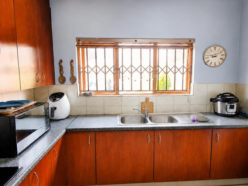 A functional kitchen featuring wooden cabinetry, laminate countertops, a stainless steel double sink, and a tiled backsplash with a window security gate.