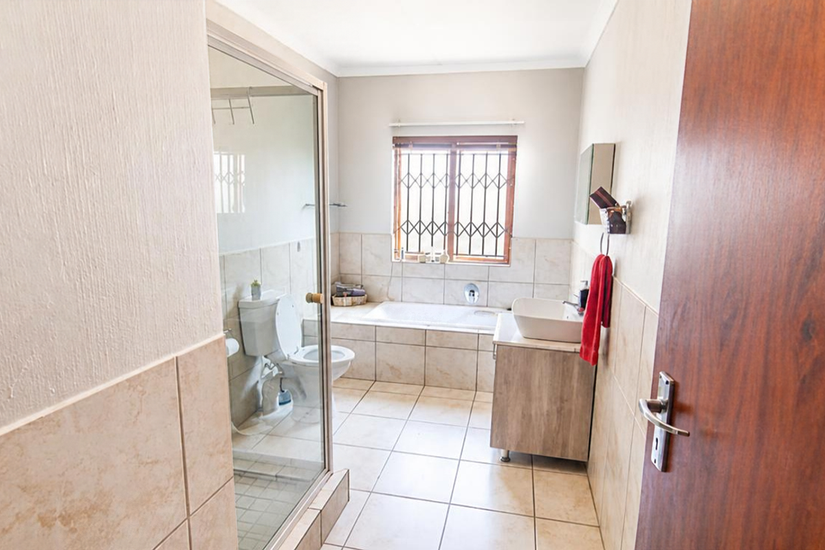 A functional bathroom featuring a built-in bathtub, a separate glass-enclosed shower, a vanity basin, and tiled flooring with security bars on the window.