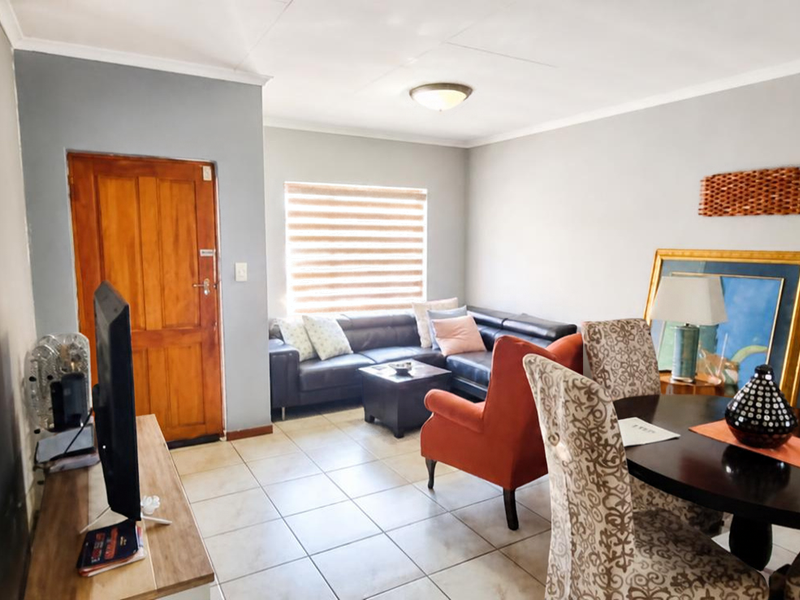 An open-plan living and dining area featuring tiled flooring, a wooden entrance door, and neutral wall finishes with natural light from a window.