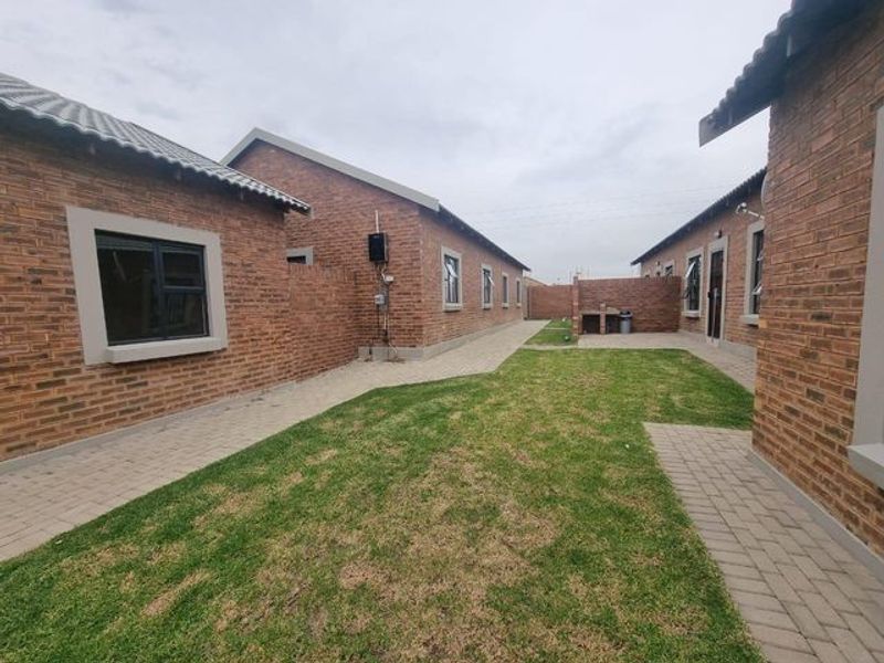 A rear exterior view of a townhouse featuring a face brick facade, paved walkways, a small lawn, and a built-in braai area.