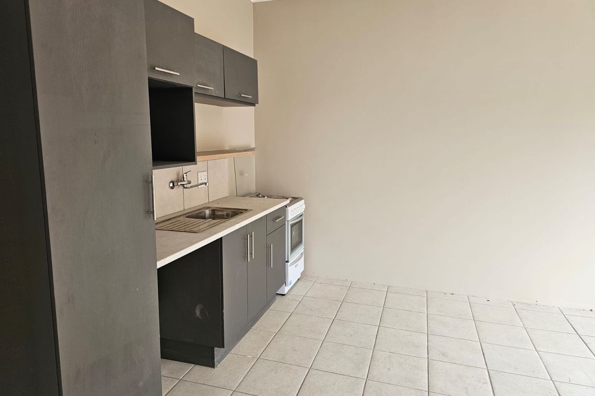 A compact, empty kitchen featuring dark grey cabinetry, a stainless steel sink, and a freestanding stove with tiled flooring.