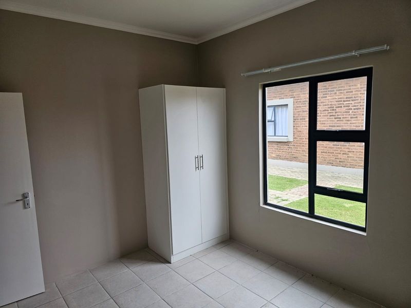 A clean, empty bedroom featuring neutral wall tones, tiled flooring, and a built-in wardrobe with a view of the exterior brick facade.