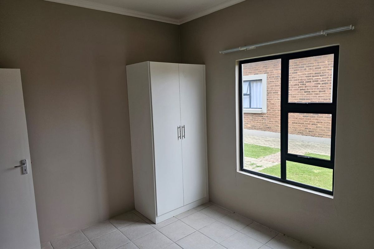 A clean, empty bedroom featuring neutral wall tones, tiled flooring, and a built-in wardrobe with a view of the exterior brick facade.