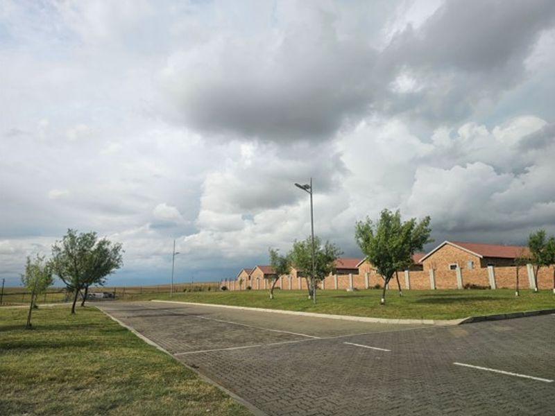 A view of the residential townhouse complex street approach, featuring paved driveways, perimeter walling, and established landscaping.