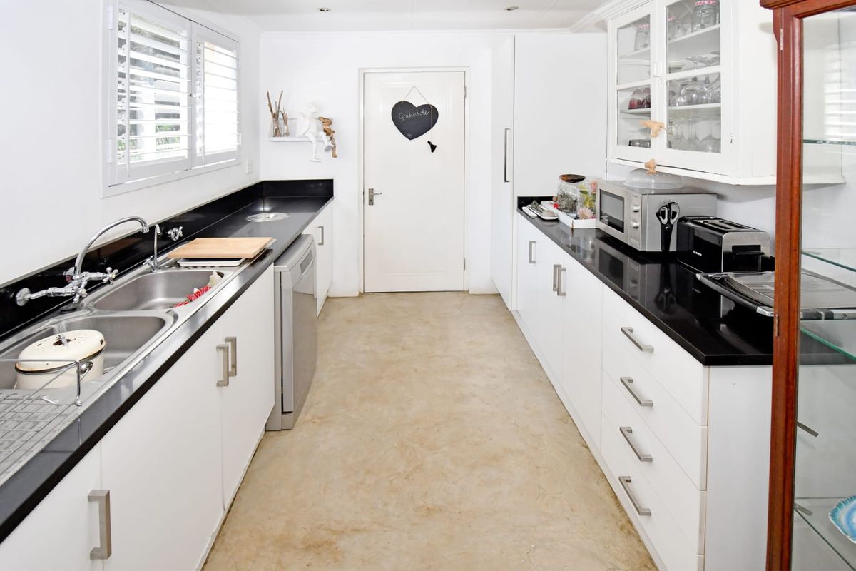 A galley-style kitchen featuring white cabinetry, black granite countertops, double basin sink, and shutter blinds, with concrete flooring throughout.
