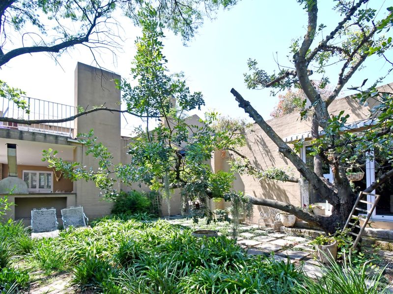 A private rear garden featuring a stone-paved patio, a built-in outdoor fireplace, and an elevated balcony overlooking the landscaped outdoor space.
