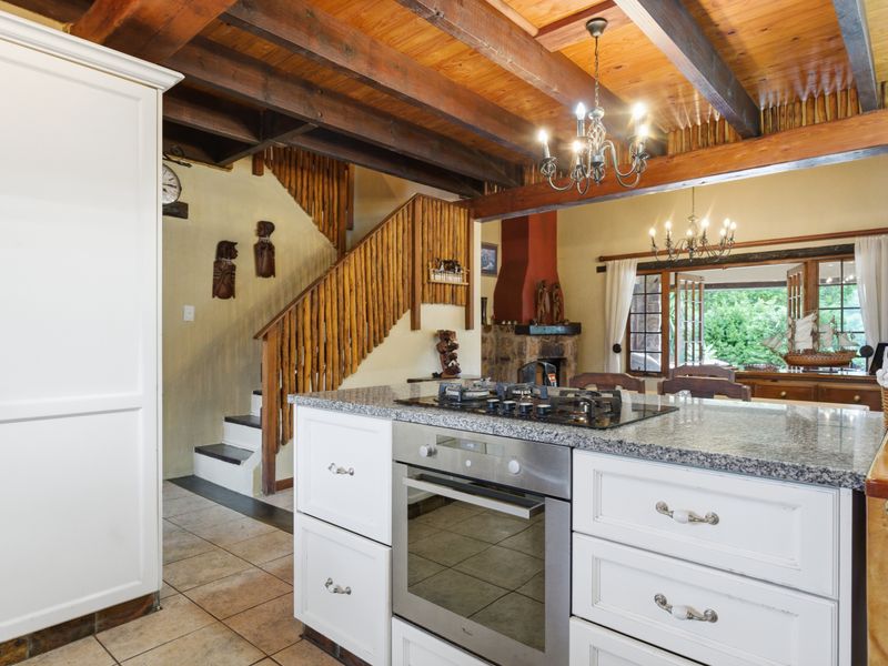 Open-plan living area featuring a kitchen island with granite countertops, exposed timber ceiling beams, a stone fireplace, and a staircase with decorative timber balustrades.