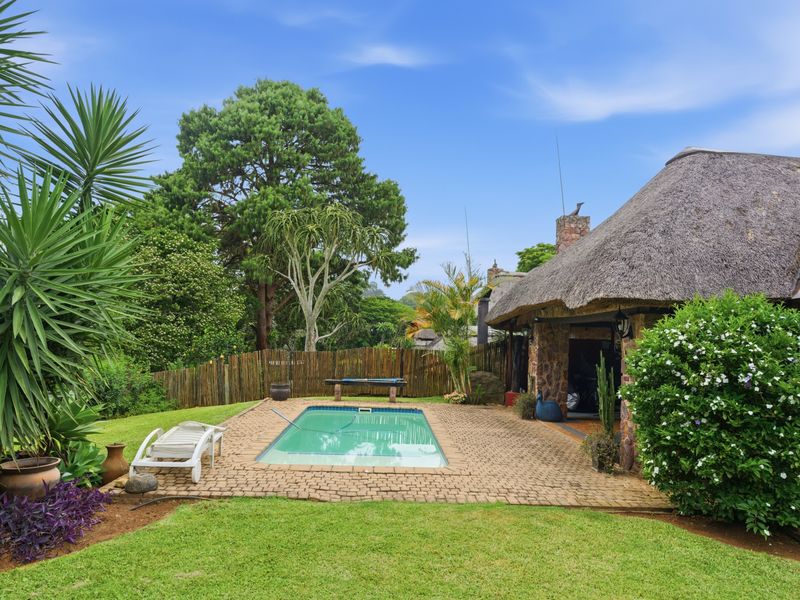 Outdoor pool area featuring a paved deck, lush garden surroundings, and a charming thatched-roof structure with stone detailing.
