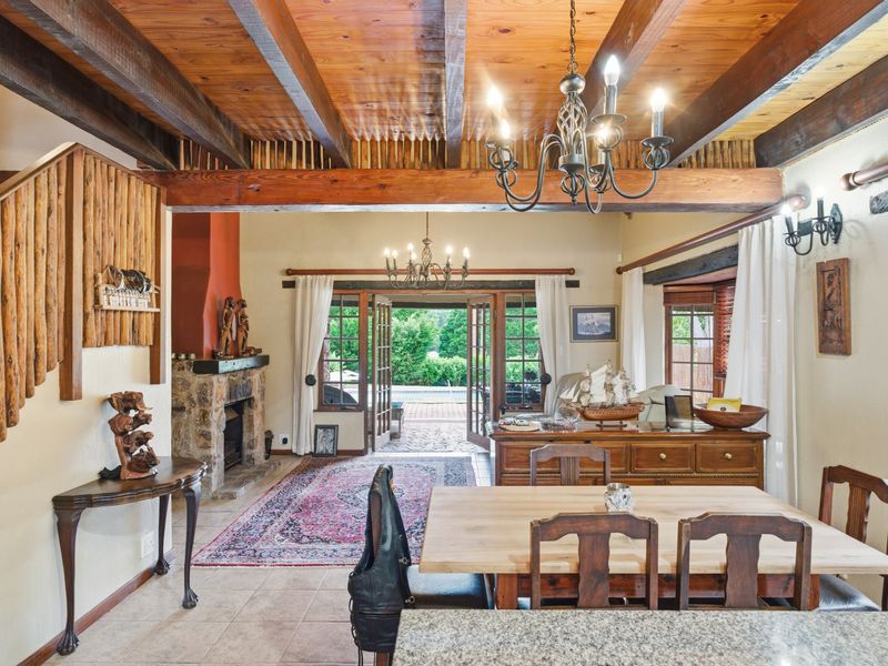 Spacious open-plan living and dining area featuring exposed timber beams, a rustic stone fireplace, and direct access to the outdoor patio through french doors.
