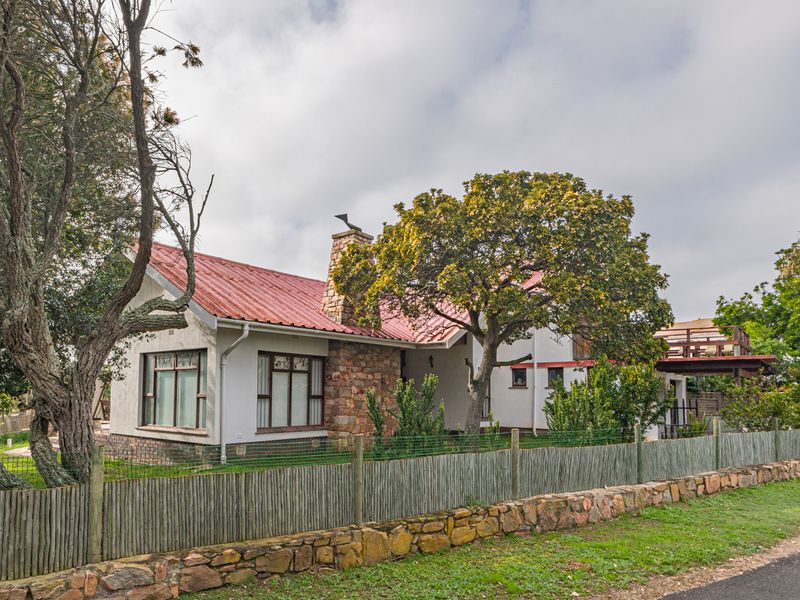 A charming single-story house featuring a red metal roof, stone chimney, and stone cladding, set behind a rustic stone and timber boundary fence.