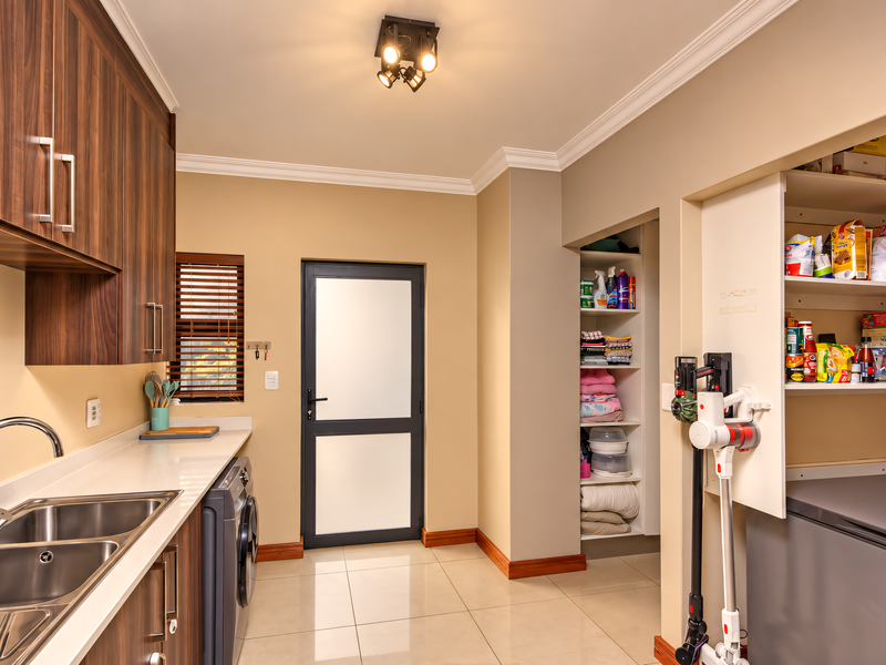 A well-organized scullery featuring modern wood-finish cabinetry, stone countertops, a double sink, and integrated shelving for ample storage and utility space.