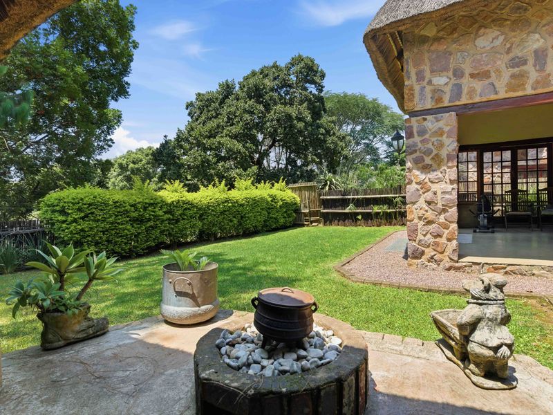 A covered patio area featuring stone cladding and a thatched roof, overlooking a well-maintained garden with a built-in fire pit.