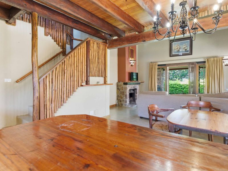 Open-plan living area featuring exposed timber beams, a unique wooden staircase, and a stone-clad fireplace, opening out to a garden view.