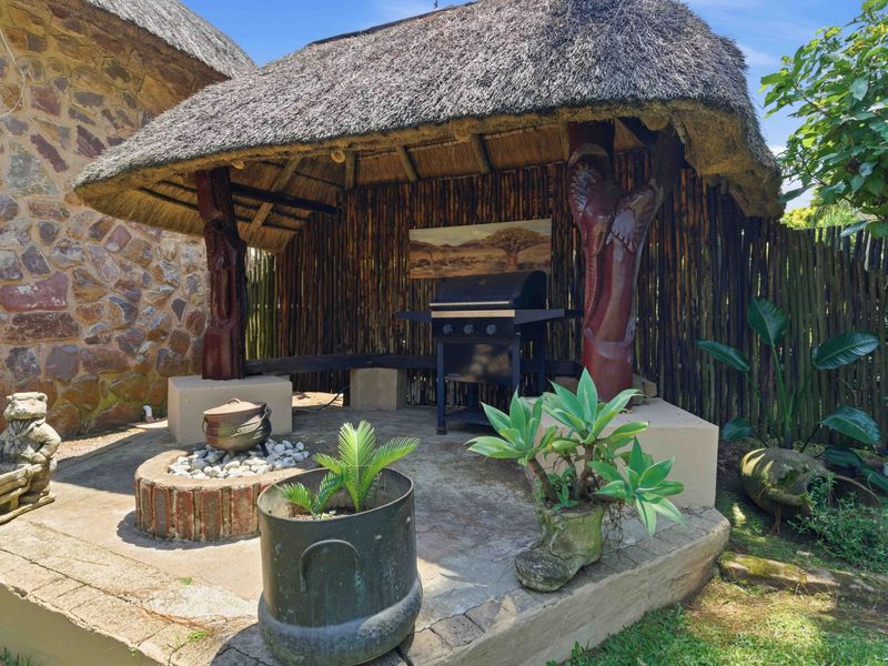 Outdoor thatched lapa area featuring a built-in fire pit and stone masonry wall, providing a rustic entertainment space in the garden.