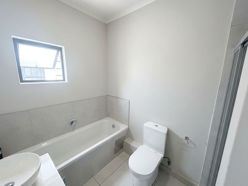 A clean, modern bathroom featuring a bathtub, toilet, and basin with neutral tiling and natural light from a top-hung window.