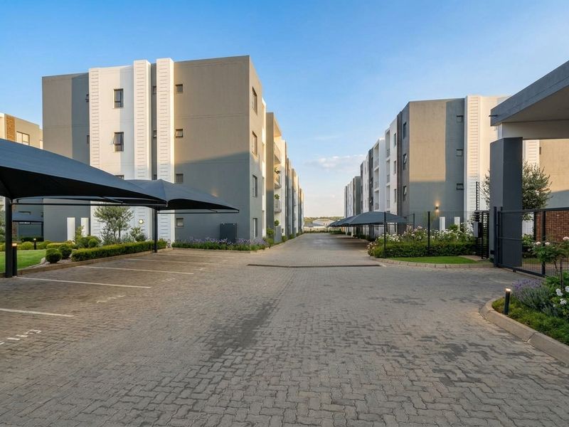 A well-maintained apartment complex entrance featuring a paved driveway, covered parking bays, and landscaped garden beds.