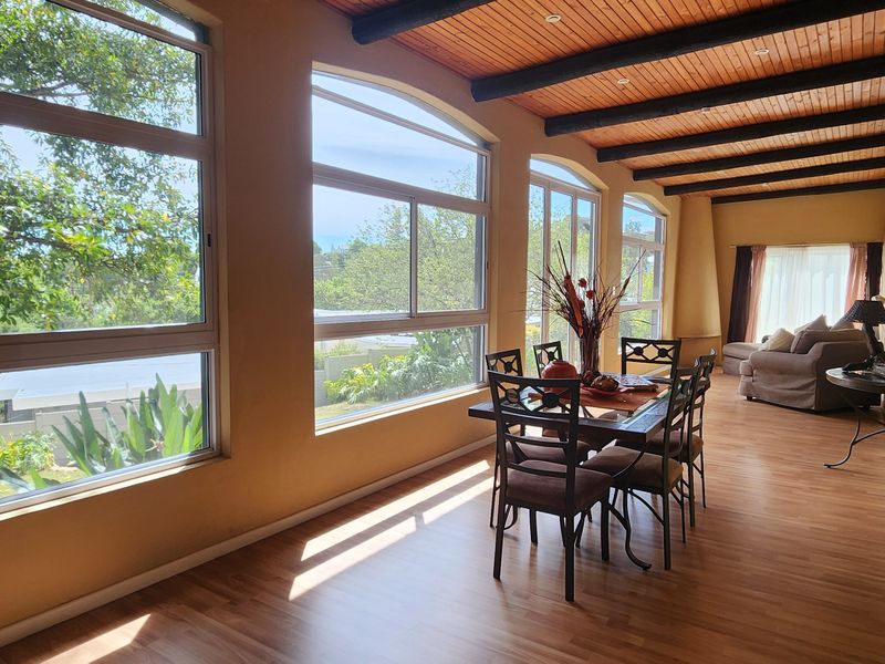 Spacious open-plan living and dining area featuring warm laminate flooring, exposed timber ceiling beams, and large windows providing ample natural light and garden views.