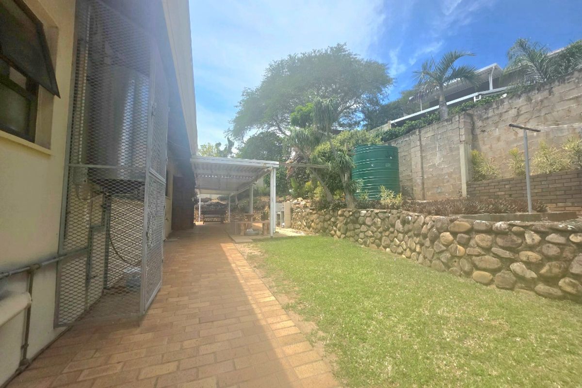 Rear exterior view featuring a paved walkway, lawn, stone retaining wall, water tank, and a covered patio area.