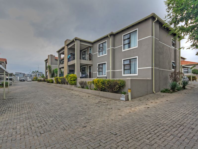 Exterior view of a multi-story apartment complex featuring paved driveway, covered parking bays, and private balconies with aluminium window frames.
